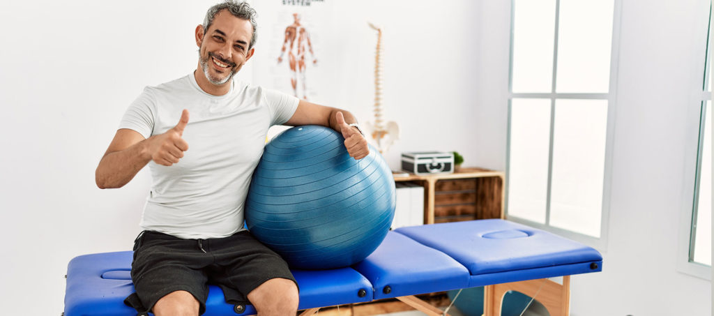 A middle aged man having a blue JOGO exercise ball, sitting on a bed, showing thumbs up after being relieved pain in JOGO Clinic