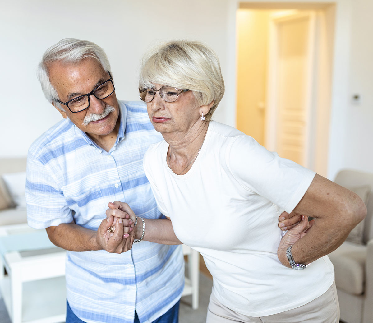 A senior old woman with back pain is supported by her husband illustrating the common risk factors for falls.